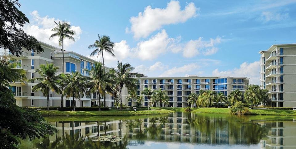 A modern apartment complex with several multi-story buildings is seen in the background. The structures feature large windows and light-colored exteriors. In the foreground, a serene pond bordered by lush green grass and leafy palm trees reflects the buildings and cloudy blue sky, reminiscent of scenes in a Phuket travel guide.