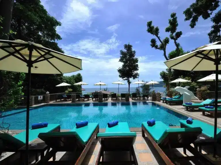 Outdoor poolside area with turquoise sun loungers, each with a blue towel. Beige umbrellas shade the loungers. The pool is clear and inviting. Trees and greenery surround the area, with clear blue skies above. In the background, there's a calm ocean view reminiscent of Patong Beach attractions.