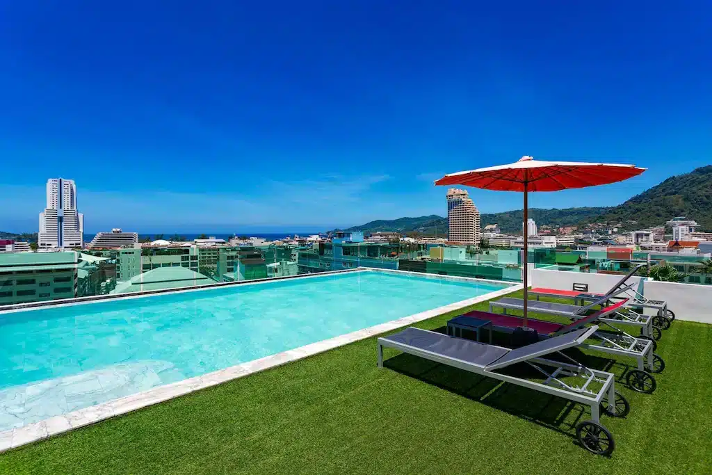 A rooftop pool with crystal clear water is surrounded by lounge chairs and a red umbrella. The space offers panoramic views of the cityscape, including tall buildings and a mountain under a clear blue sky. The ground around the pool is covered in green turf, making it one of the best places to visit in Patong.