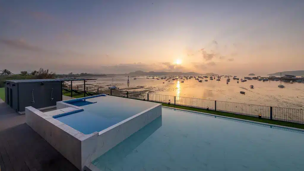 A serene coastal scene at sunrise features an infinity pool in the foreground, with a smaller elevated pool beside it. The calm sea stretches out in the background, scattered with boats. Wat Chalong Phuket rises over distant hills, casting a warm glow over the water and tinged sky of dawn.