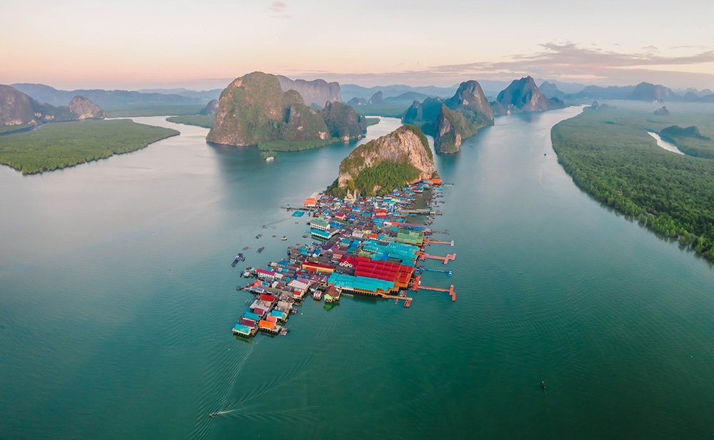 Aerial view of a picturesque island with lush green hills surrounded by calm waters. Vibrantly colored buildings are clustered on the island, with green forests extending from the shore. The horizon shows more green hills and distant mountains under a soft, pastel sky at dawn or dusk—a perfect scene for any Phuket travel guide.