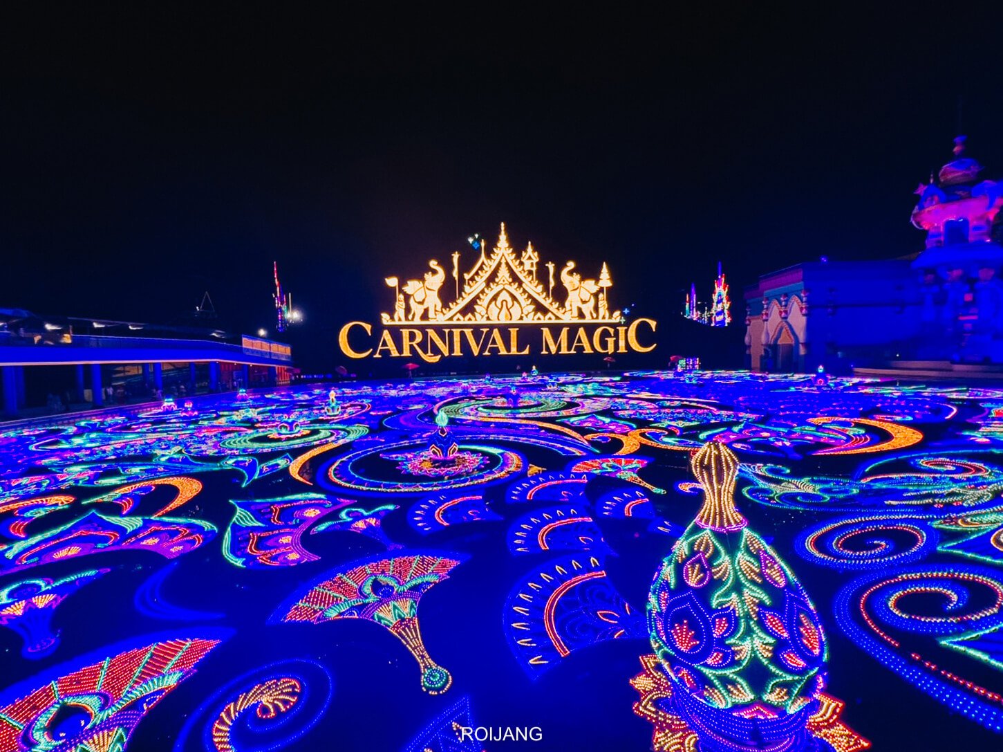 A vibrant nighttime scene of "Carnival Magic" features a large illuminated sign with ornate golden details. The foreground is adorned with intricate, colorful neon light designs forming patterns on the ground, including a prominent peacock figure, creating a dazzling display—a must-see in any Phuket travel guide.