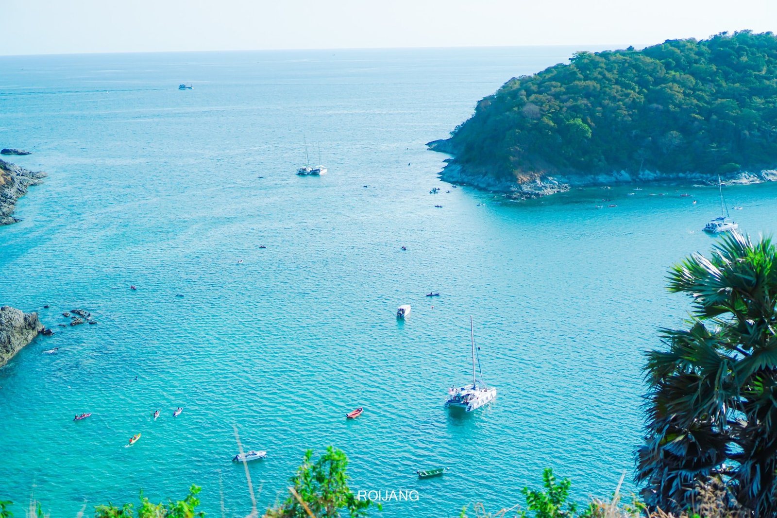 An aerial view of a serene coastal scene featured in many Phuket travel guides, showcasing turquoise waters, several anchored sailboats, and small boats dotting the bay. A lush, forested peninsula extends into the water, with rocky outcrops and sparse greenery lining the shore, including a visible palm tree in the foreground.