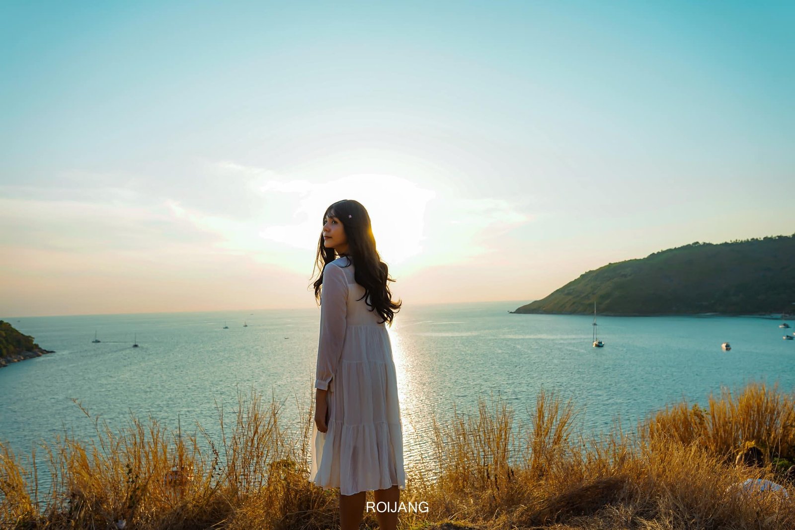 A woman in a flowing white dress stands on a grassy cliff at sunset, overlooking a tranquil sea with a clear sky. The sun is setting behind her, casting a golden glow. Sailboats are anchored in the distance near lush, green hills that frame the serene ocean view—reminiscent of Phuket attractions highlighted in any Phuket travel guide.