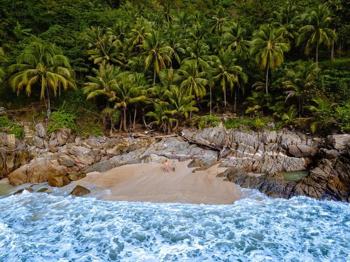 An aerial view of a small, secluded beach surrounded by rocky terrain and lush green palm trees. Two people are on the sandy shore near the edge of the forest. Waves from the blue ocean are crashing onto the beach. This tropical, picturesque setting is a must-see in any Phuket travel guide.