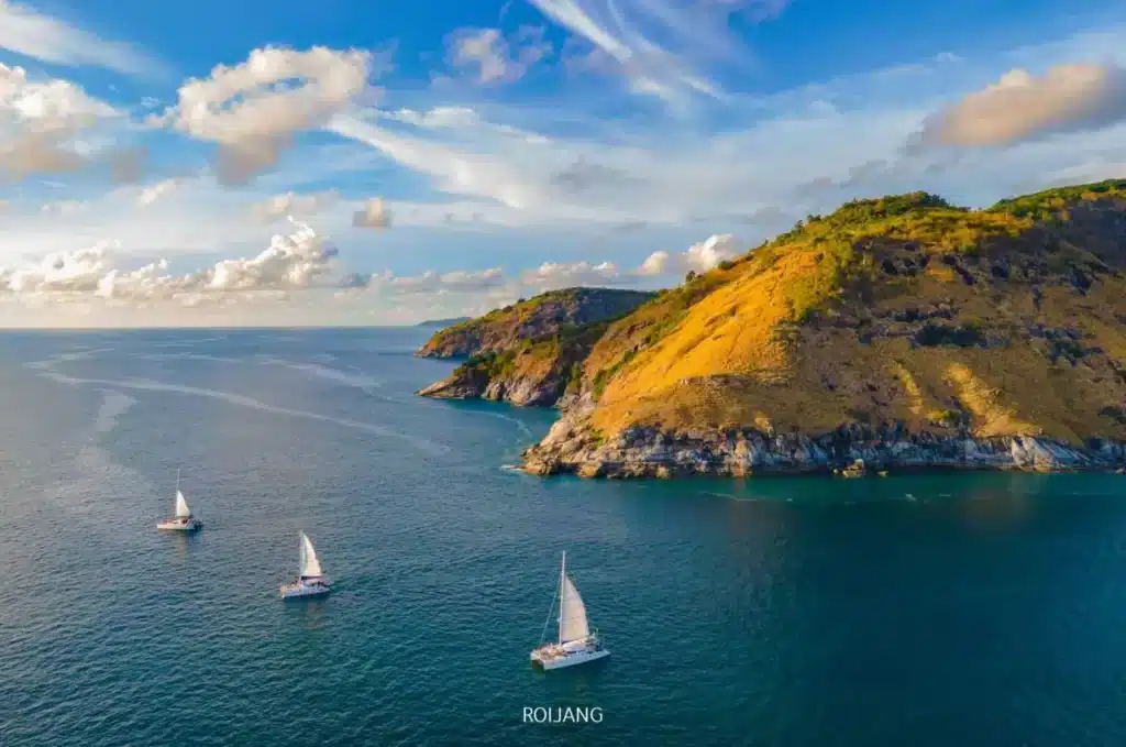 A vibrant coastal scene shows four white sailboats gliding across the blue ocean near a rocky coastline. The lush, green hills are bathed in golden sunlight, reminiscent of the serene landscape surrounding Wat Chalong Phuket. The sky above is dotted with white clouds as the horizon stretches out under wispy blues.