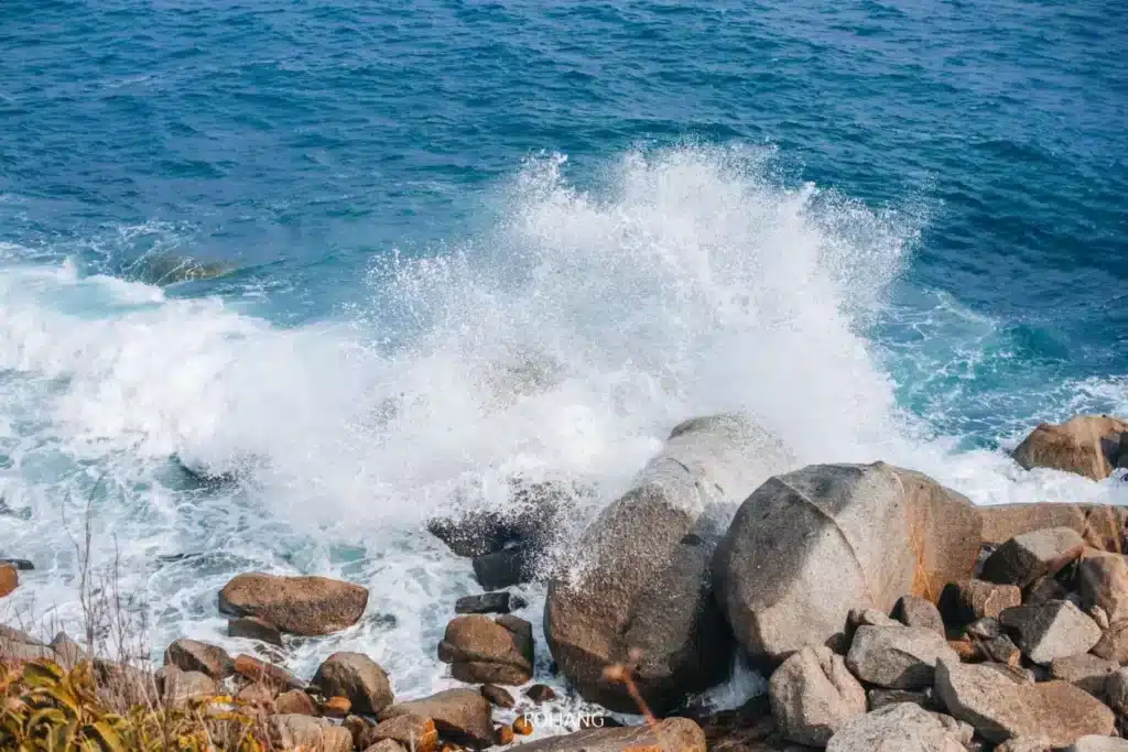 Waves crash against large rocks on a rocky coastline under a bright blue sky, reminiscent of the serene backdrop near Wat Chalong Phuket. The ocean water is vivid blue, with white foam and spray from the impact. The scene is framed by scattered boulders and some dry vegetation in the foreground.