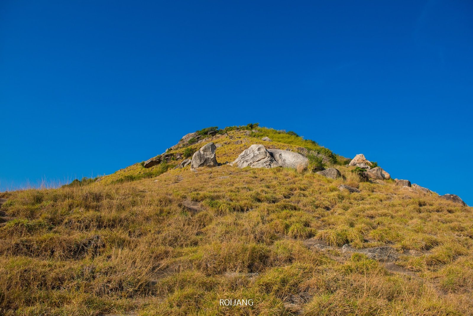 A serene, grassy hill with scattered rocks under a clear, deep blue sky. Covered in dry, golden-brown grass and sparse green vegetation, the hill stands out as a natural gem. Prominent large rocks near the top add texture to this scene reminiscent of serene Phuket attractions.