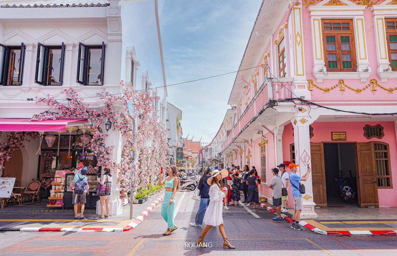 Pedestrians walk through a vibrant street lined with colorful buildings in pastel shades, including white and pink. One building is adorned with cherry blossom decorations. People are taking photos, chatting, and sightseeing. The sky is clear with some white clouds, creating a bright and cheerful atmosphere—a perfect snapshot for any Phuket travel guide.