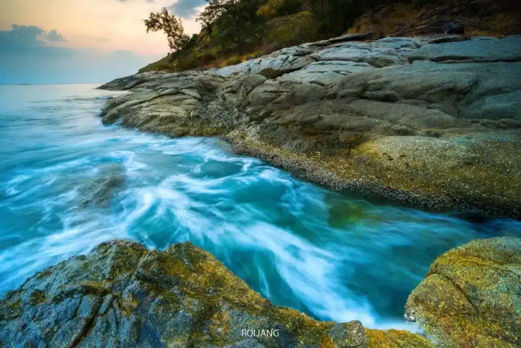 A tranquil coastal scene during sunset with rugged rock formations and turbulent white water flowing between them into a serene, calm turquoise sea. Overhanging trees add to the picturesque nature of this landscape, reminiscent of the peaceful ambiance around Wat Chalong Phuket.