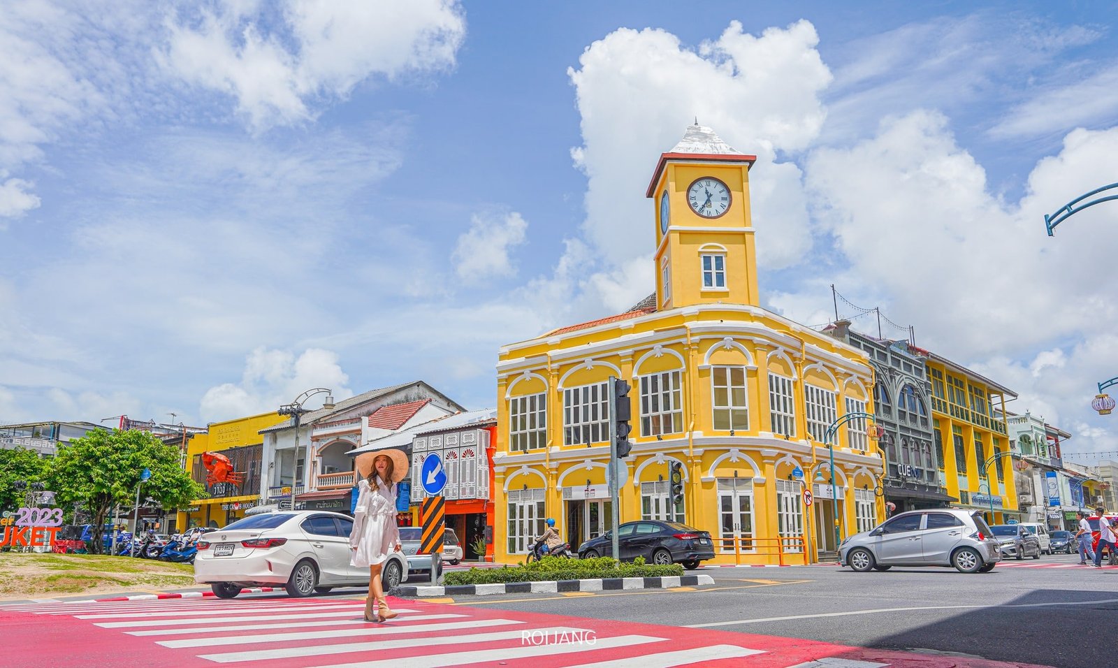 A woman in a white dress and wide-brimmed hat walks across a red and white striped crosswalk. Behind her is a vibrant yellow colonial-style building with a clock tower, one of the highlights in any Phuket travel guide. The sky is bright blue with white clouds, and colorful facades surround the scene.