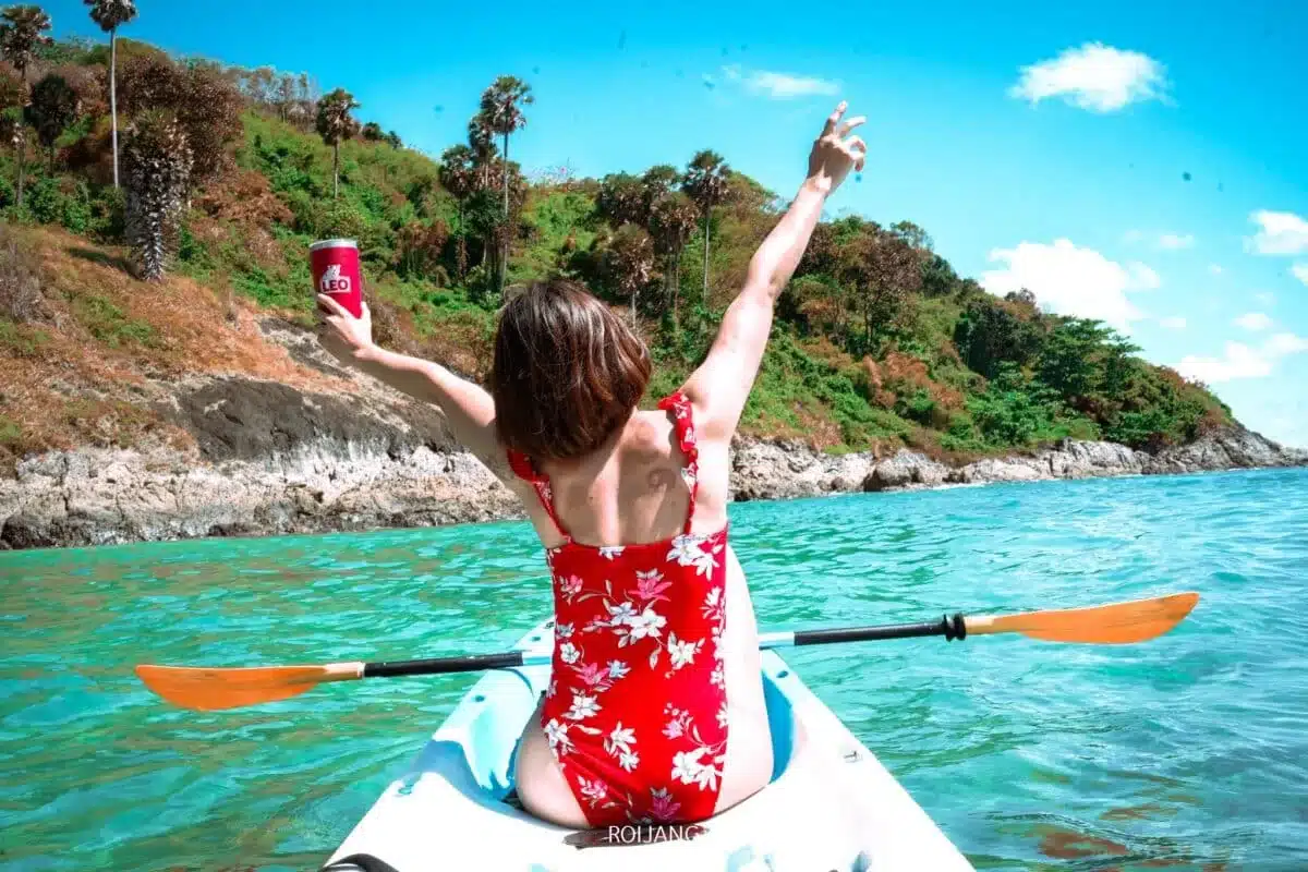 A person in a red and white floral swimsuit raises their arms in joy while seated in a kayak on turquoise waters near a rocky shoreline with lush greenery. They hold a red beverage can in one hand. Enjoying one of the best places to visit in Patong, the bright blue sky has few clouds above.