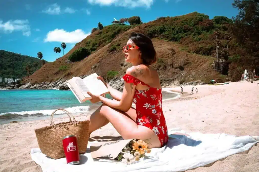 A woman in a red floral swimsuit sits on a white blanket at a sandy beach near Laem Phromthep, reading a book. She wears sunglasses and has a tattoo on her arm. By her side are a straw bag, a stainless steel tumbler, and a small bouquet of flowers. The background features rolling hills, palm trees, and clear blue skies.