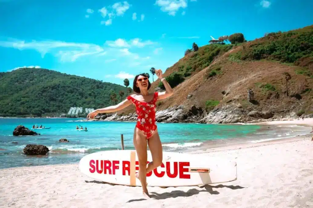 A woman in a red floral swimsuit is joyfully jumping on a sunny beach at Laem Phromthep. Behind her, a surf rescue board is placed on the sand. The background features clear turquoise water, swimmers, and lush green hills under a bright blue sky with scattered clouds.