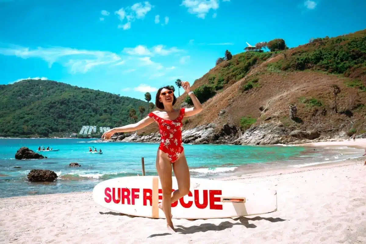 A woman in a red floral swimsuit is joyfully jumping on a sunny beach with turquoise water and green hills in the background. Near her stands a white surf rescue board with red lettering, while several people swim nearby. Enjoy moments like these when exploring the best places to visit in Patong.