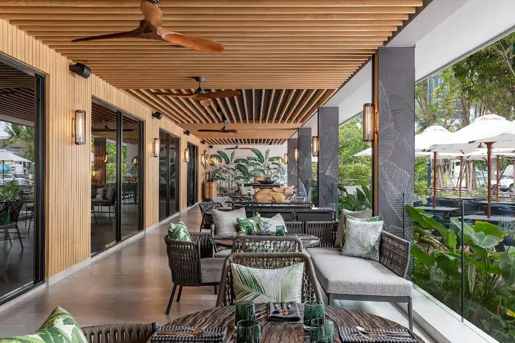 Outdoor restaurant seating area with modern wooden ceiling, large windows, and ceiling fans. Tables are set with green glassware and patterned cushions adorn the chairs and benches. Plants and trees surround the open-air space, with white umbrellas visible in the background—one of the best places to visit in Patong.