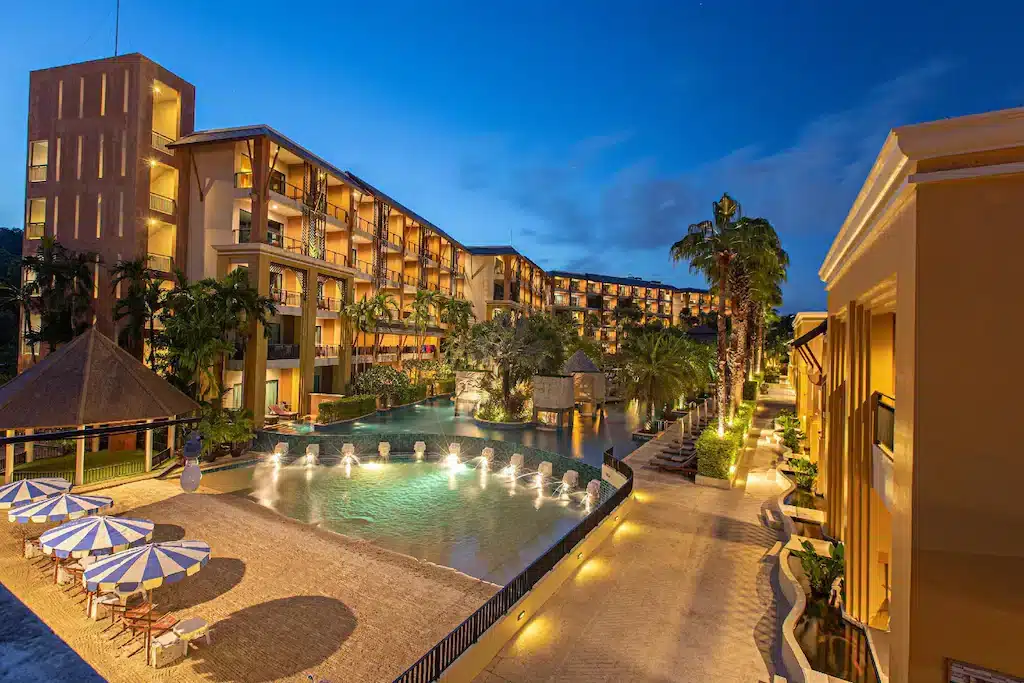 A tropical resort at dusk featuring an illuminated swimming pool with several water fountains. Surrounding the pool are sun loungers, striped umbrellas, and tall palm trees. In the background, multi-story buildings with balconies overlook the pool, reminiscent of the serene ambiance at Laem Phromthep Sunset Viewpoint.