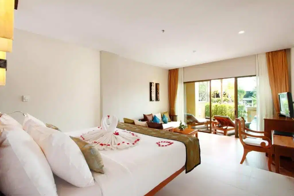         A spacious hotel room with a large bed adorned with a towel art swan and red flower petals. There's a seating area with a sofa and chairs, a flat-screen TV on a wooden desk, and large windows with golden curtains that lead to a balcony. From there, enjoy the breathtaking Laem Phromthep Sunset Viewpoint.