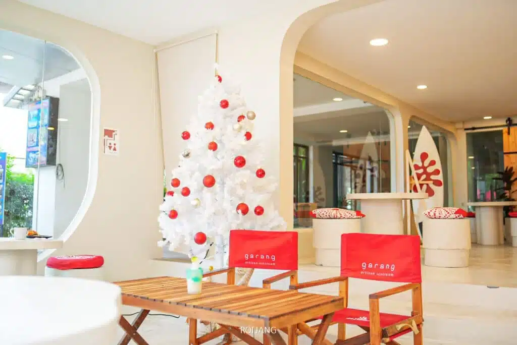 A cozy indoor cafe with white walls featuring festive holiday decor. A white Christmas tree adorned with red and gold ornaments stands prominently. Red chairs with the word "garang" face a wooden table. Modern indoor furniture and decor, inspired by Laem Phromthep's serene ambiance, contribute to the welcoming atmosphere.