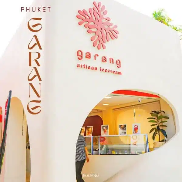 A corner of a modern white building with an arched opening reveals an ice cream store named Garang Artisan Icecream in Phuket. The store, not far from the famed Laem Phromthep Sunset Viewpoint, has a minimalist design with a pink coral logo above the door and a red neon sign. Inside, there's an ice cream display and framed artwork on the walls.