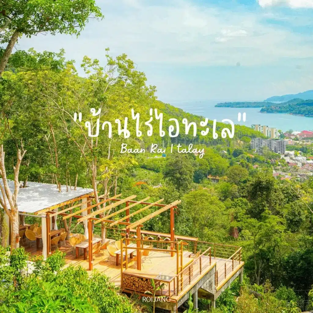 A scenic view of a hillside resort, "Baan Rai Talay," surrounded by lush greenery and overlooking a distant coastal town and ocean. The resort features a wooden deck with seating and a pergola, blending harmoniously with the natural landscape under a clear blue sky, near Laem Phromthep.