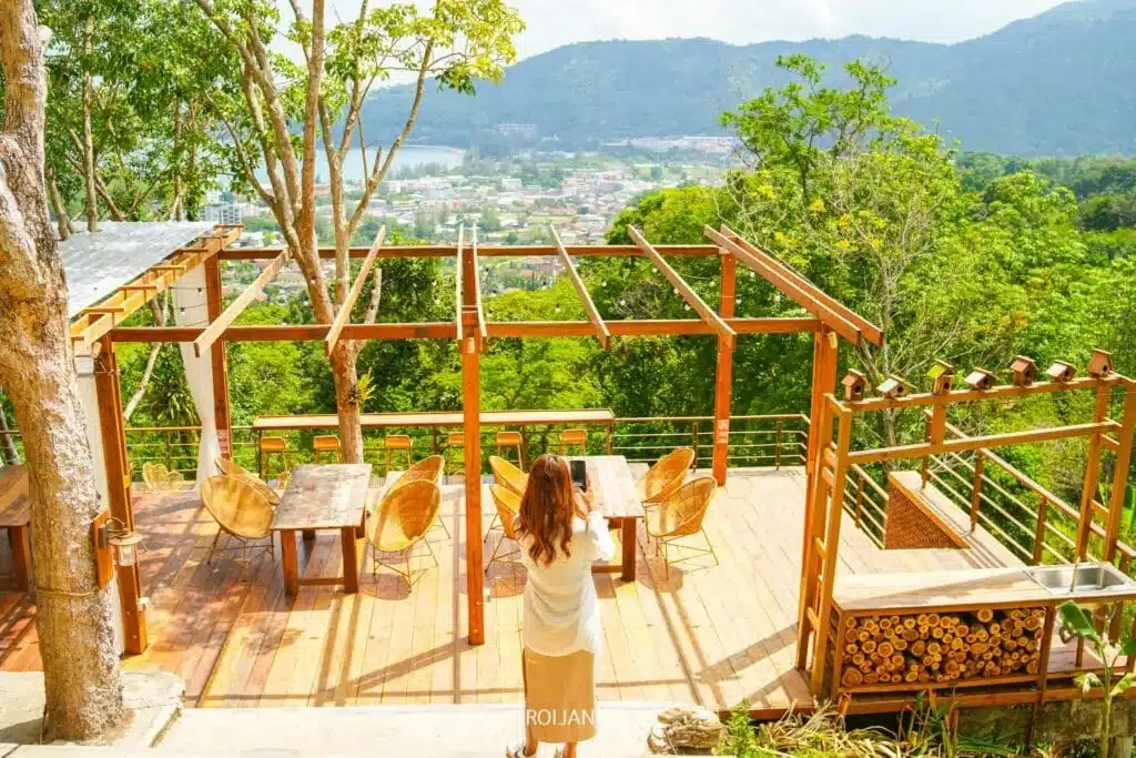 A person with long hair and a light-colored dress stands on a wooden deck with rustic furniture, looking out at the scenic Laem Phromthep Sunset Viewpoint. The deck is surrounded by lush greenery and overlooks a valley and distant town, with mountains in the background under a clear sky.