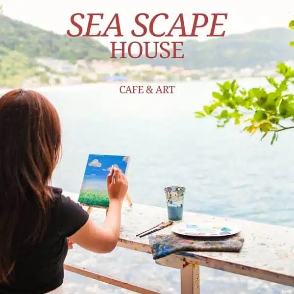 A person with long hair is painting a seascape on a canvas at Laem Phromthep, a picturesque seaside location. Art supplies, including brushes, a paint palette, and a water container, are on a ledge in front of them. "SEA SCAPE HOUSE CAFE & ART" is written at the top of the image.