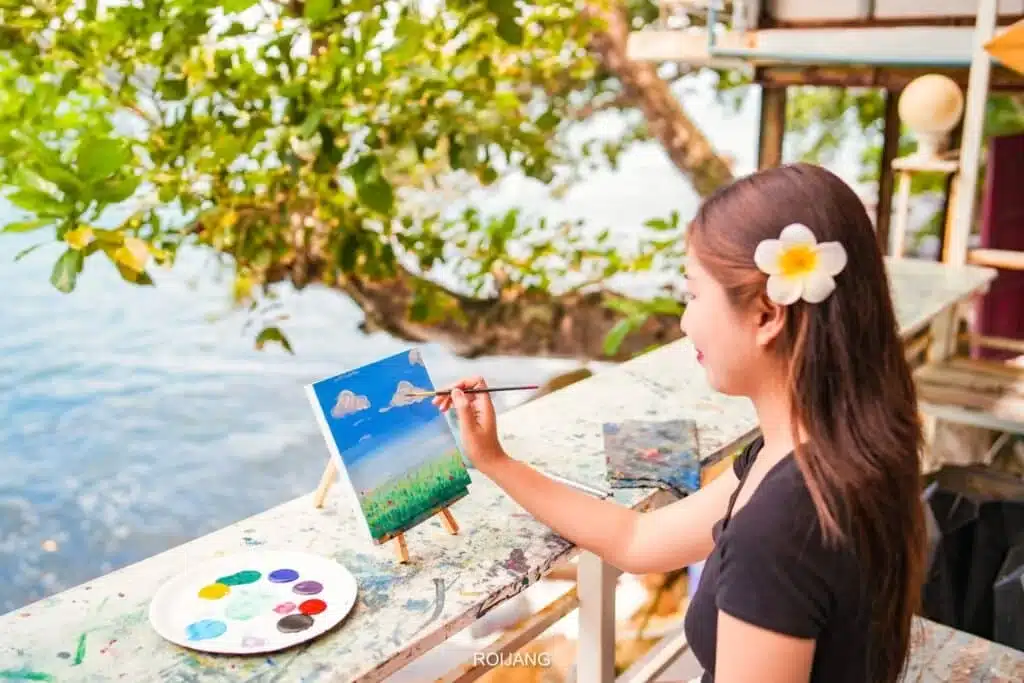 A woman with long hair and a white flower tucked behind her ear is painting a landscape on a small canvas. Seated at an outdoor table near a tree, with the serene beauty of Laem Phromthep Sunset Viewpoint in the background, she has various colored paints arranged on a palette beside her.