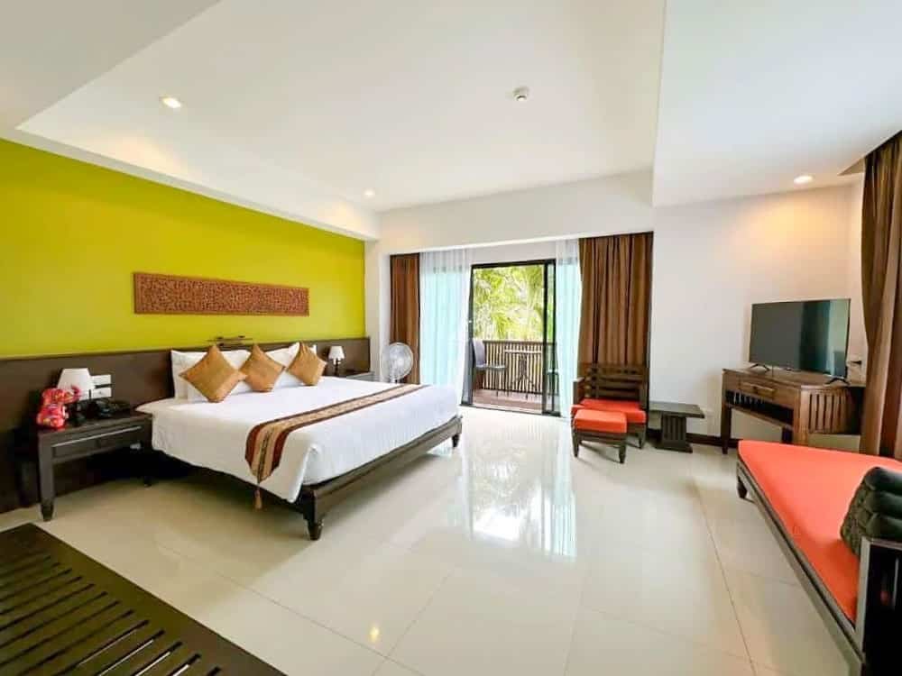 A spacious hotel room with a white-tiled floor, featuring a large bed with white linens and brown and orange accent pillows against a bright green accent wall. The room includes a television, a lounge chair, a small table, and a balcony with views of greenery reminiscent of the Laem Phromthep Sunset Viewpoint.