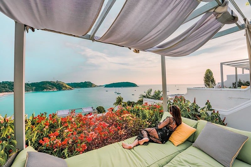 A person reclines on a green daybed under a white canopy, overlooking the scenic Laem Phromthep Sunset Viewpoint. The foreground features vibrant red flowers and lush green foliage. The background includes a calm blue sea with anchored boats and a hilly shoreline under a clear sky.