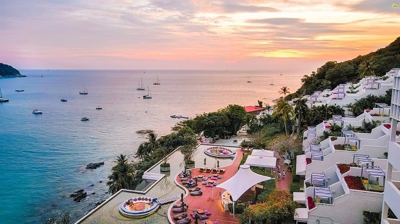 Aerial view of a coastal resort at sunset near Laem Phromthep, featuring terraced white buildings with balconies descending a hillside. The resort has vibrant landscaping, including pools and pathways. The ocean extends to the horizon with several anchored boats, and the sky displays shades of orange and pink.