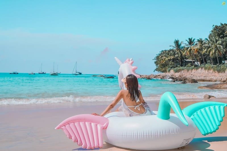 A person in a white swimsuit relaxes on an inflatable unicorn float in shallow, clear blue water near a sandy beach. Palm trees and boats on the water are visible in the background under a bright blue sky, making it feel like a scene straight out of a Phuket travel guide.