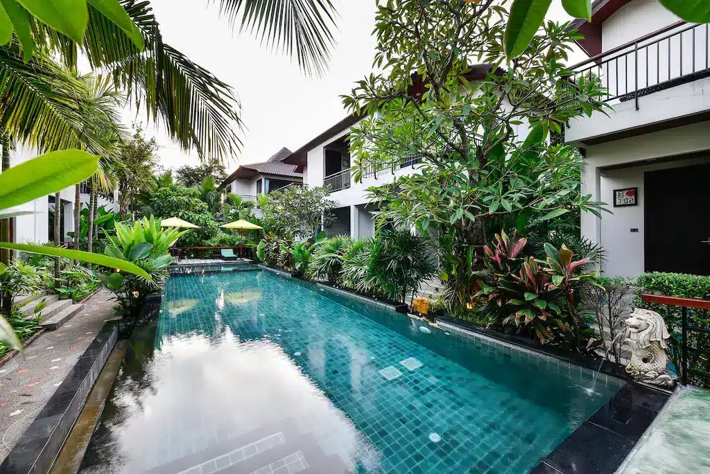 A serene outdoor pool is surrounded by lush tropical plants and trees. The pool, with clear blue water, is flanked by modern two-story white buildings with balconies. Steps lead to the pool, with a stone statue on the right. Green umbrellas and lounge chairs are visible in the background near Chalong Temple.