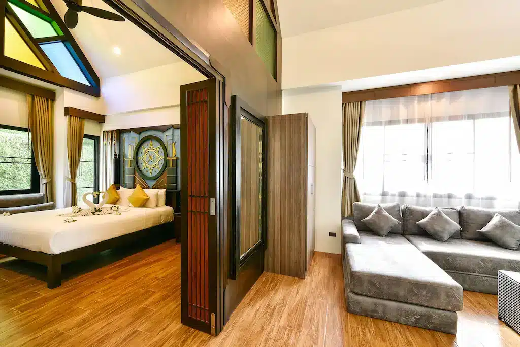 A spacious, modern hotel suite with a bedroom and living area. The bedroom features a large bed with white linens and yellow pillows, framed by a stained glass window. The living area has a gray sectional sofa and large windows with sheer curtains, allowing ample natural light. Wood flooring throughout, echoing the serene ambiance of Wat Chalong Phuket.