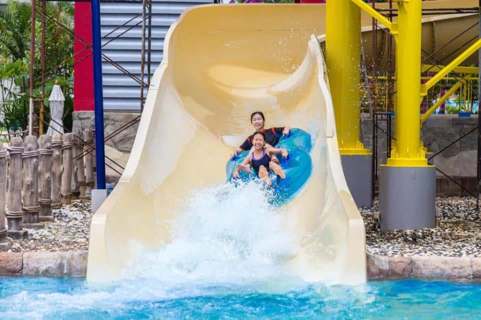 Two people, one adult and one child, joyfully slide down a large yellow water slide at a colorful Phuket attractions water park. They are riding a blue inflatable ring with splashes of water at the bottom, surrounded by vibrant structures and lush greenery in the background.