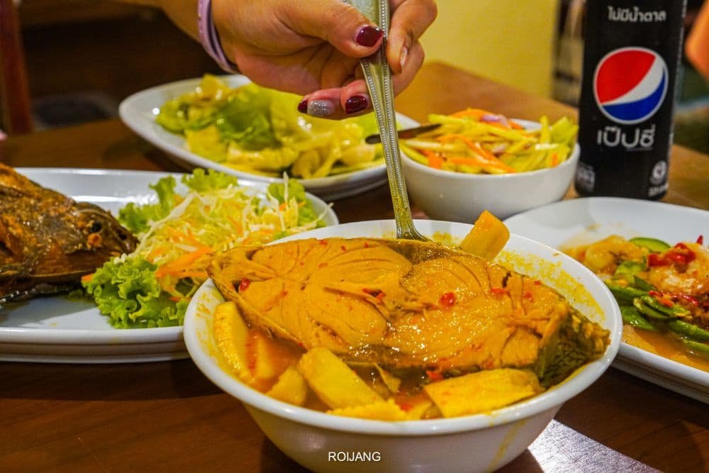 A hand with painted nails holds a spoon above a bowl of yellow curry with fish and bamboo shoots in a Phuket travel guide-worthy restaurant. Surrounding dishes include a whole fried fish on a bed of lettuce, sliced vegetables, and a black can of Pepsi—all set elegantly on a wooden table.