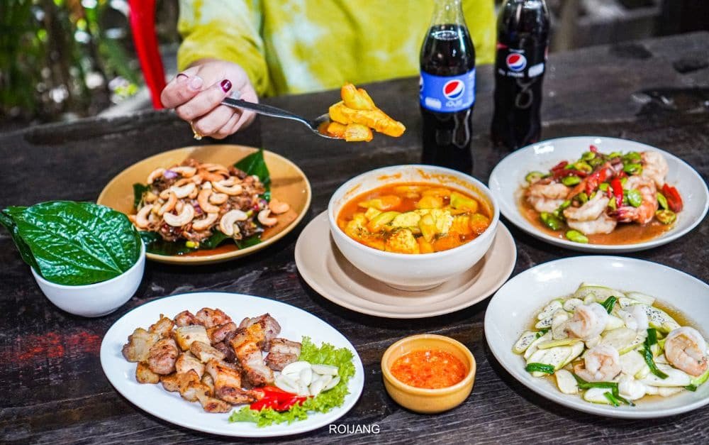 A table set with various Thai dishes, including a hand holding a spoonful of yellow curry. Dishes include a plate of nuts and greens, orange curry, grilled meat with dipping sauce, shrimp and vegetables, and shrimp with cucumber. Two bottles of Pepsi in the background nod to delights found in any Phuket travel guide.