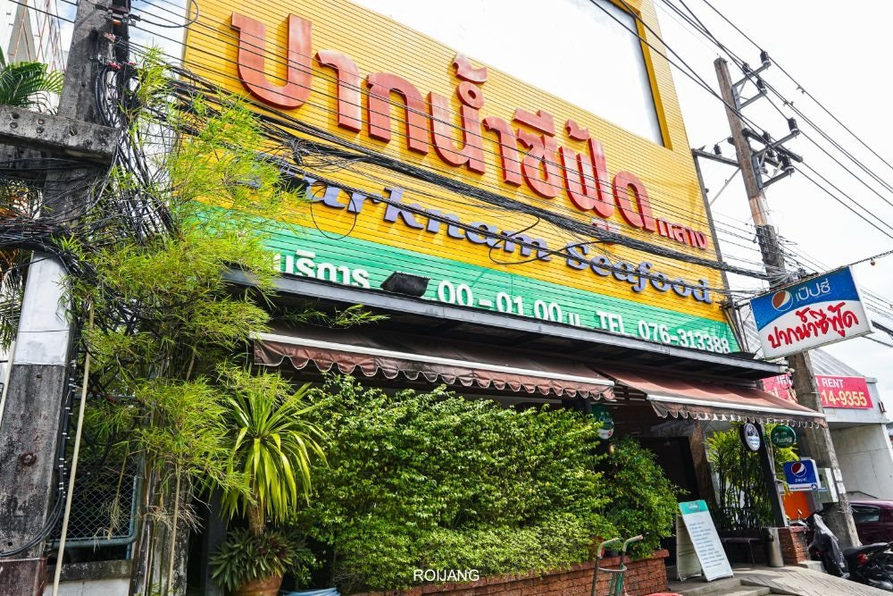 Shopfront featuring a vibrant yellow banner with large red and green Thai script and "Mahanakorn Seafood" in English. Beneath the banner, lush foliage and a small awning accentuate the scene. Power lines and a weathered utility pole are visible to the left, along with nearby Thai signage—a true gem in any Phuket travel guide.