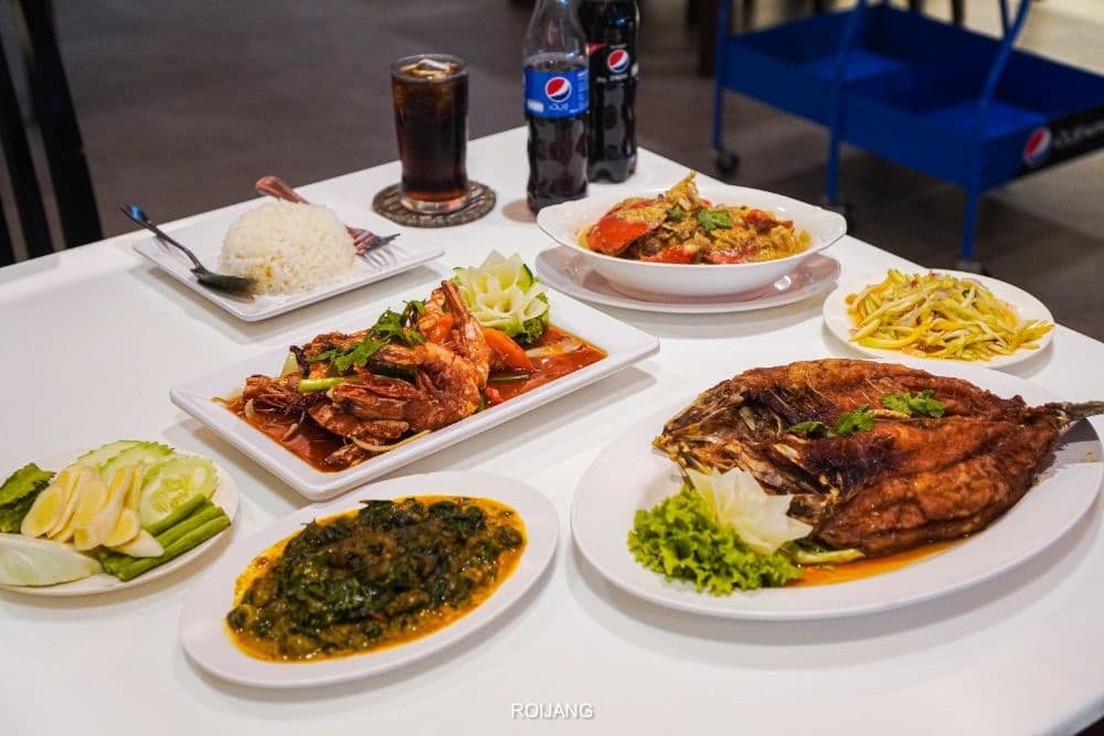 A table with various Thai dishes, reminiscent of a Phuket travel guide, features a whole fried fish garnished with greens, stir-fried shrimp with vegetables in sauce, a bowl of greens in yellow curry, sliced cucumbers and lime, rice, another seafood dish, and a bottle and glass of Pepsi.