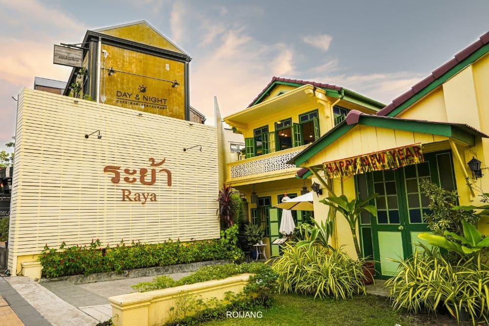 A charming scene from a cozy Thai resort, known for being one of the top Phuket attractions. The image features a two-story yellow building with green doors and trim, adorned with a "Happy New Year" sign. To the left, a white wall with the word "Raya" stands near another building labeled "Day & Night." Lush greenery surrounds the area.