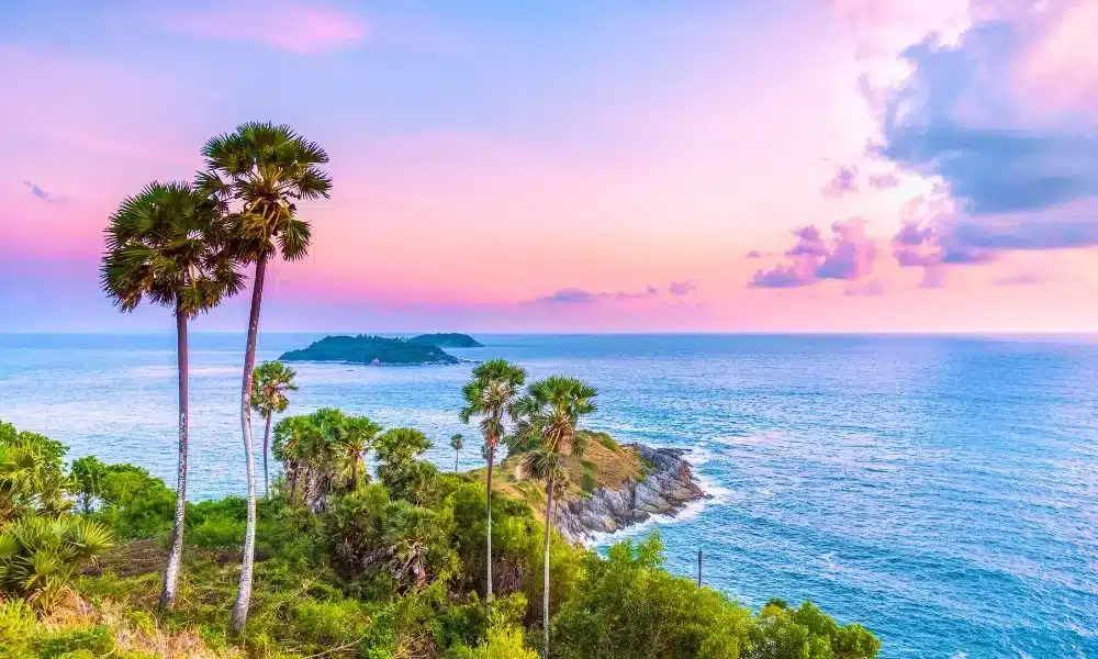 A vibrant coastal scene at sunset with a colorful sky transitioning from pink to blue. Tall palm trees and lush greenery frame the foreground, leading to a rocky peninsula jutting into the calm ocean. Laem Phromthep Sunset Viewpoint offers a breathtaking view of a small, distant island on the horizon under a partly cloudy sky.