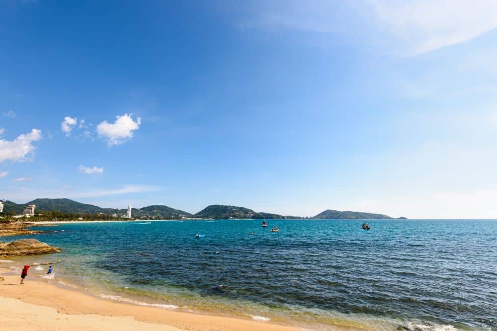 A beach scene showcasing a sandy shoreline with a few people near the water's edge. The calm, blue ocean stretches out to distant, tree-covered hills under a clear, sunny sky. Boats float on the water, and white, fluffy clouds dot the sky—truly one of the best places to visit in Patong.