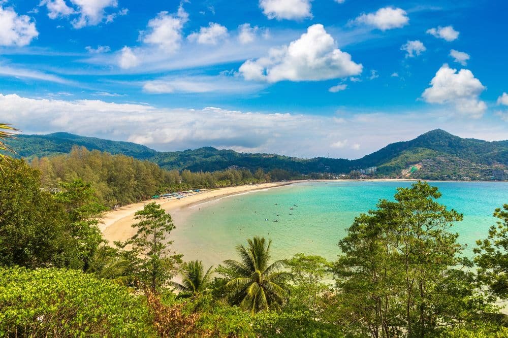 A scenic view of Patong Beach with clear blue skies and scattered clouds. This tropical paradise features golden sand bordered by lush green hills and trees. The turquoise ocean water is calm, with a few boats visible in the distance, while palms and other foliage frame the foreground.