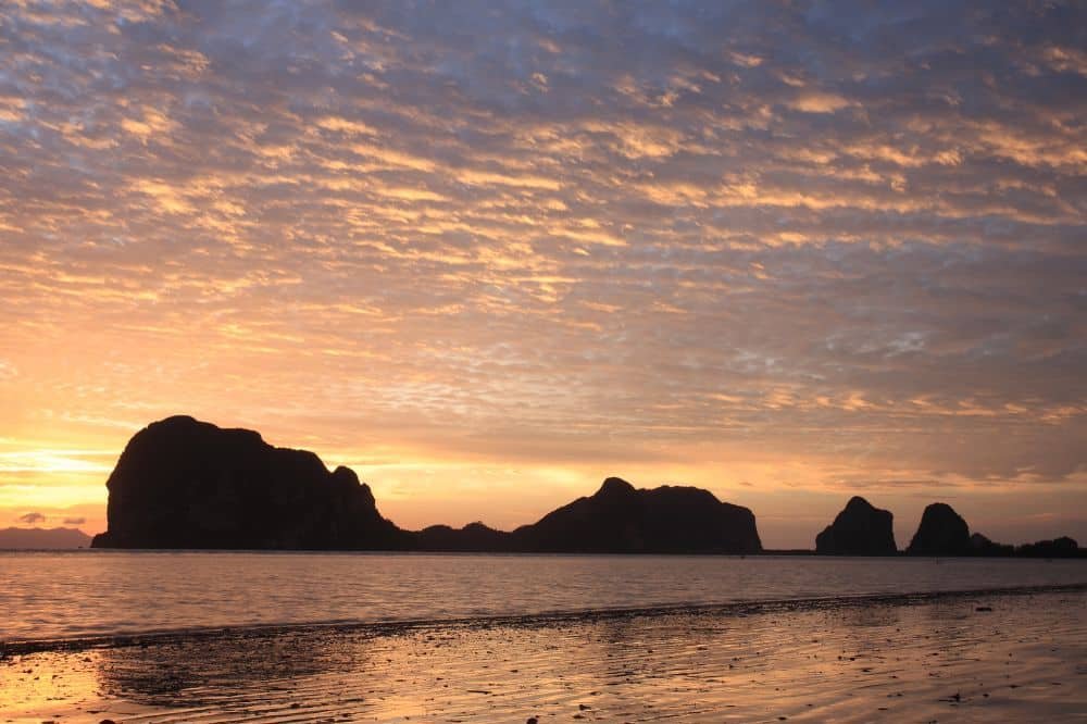 A serene beach at sunset with calm waters reflecting the vibrant orange, pink, and purple hues of the sky. Several dark rock formations and small islands are silhouetted against the horizon, adding to the tranquil and picturesque scene. The beach is deserted, enhancing the feeling of peace—a stark contrast to the lively Patong nightlife nearby.