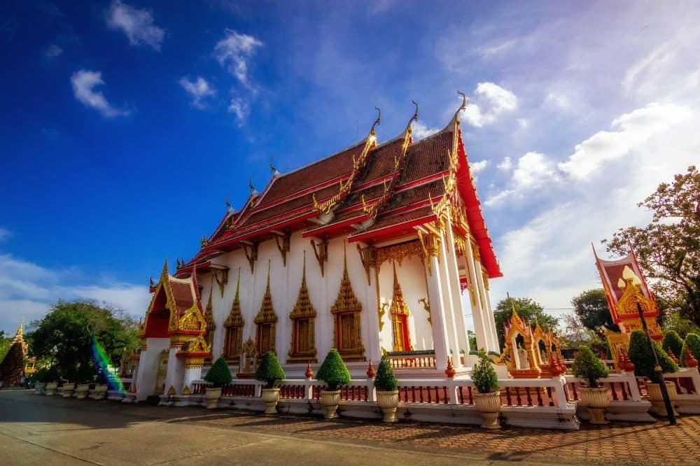 A grand Thai temple, known as Wat Chalong Phuket, with intricate gold details and an ornate red roof stands majestically under a bright blue sky with scattered clouds. Small manicured shrubs line the pathway in front of Chalong Temple, while smaller structures and trees are visible in the background.