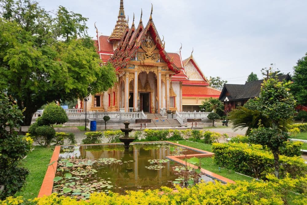 A vibrant Buddhist temple in Thailand, known as Wat Chalong Phuket, with ornate golden details and a red roof is surrounded by lush greenery. In the foreground, a calm pond with water lilies is bordered by yellow flowers and neatly trimmed bushes. The sky above is overcast, giving the scene a serene ambiance.