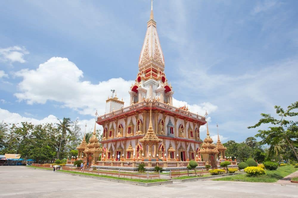 A Thai temple with a tall, ornate spire stands against a blue sky with a few clouds. Wat Chalong Phuket features intricate gold and red details, multi-tiered roofs, and golden statues. The surrounding area has manicured greenery, small trees, and paved pathways.