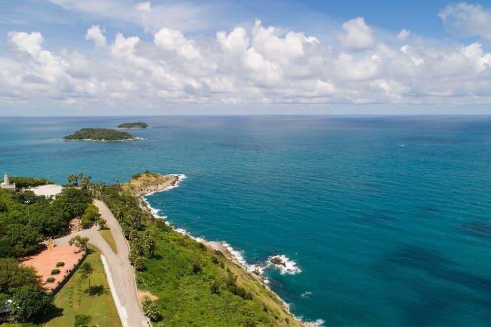 Aerial view of Laem Phromthep coastline with a winding road on the left bordered by lush greenery. The blue ocean extends to the horizon, with two small islands visible in the distance under a partly cloudy sky. The shoreline features rocky outcrops and gentle waves, perfect for a Laem Phromthep sunset.