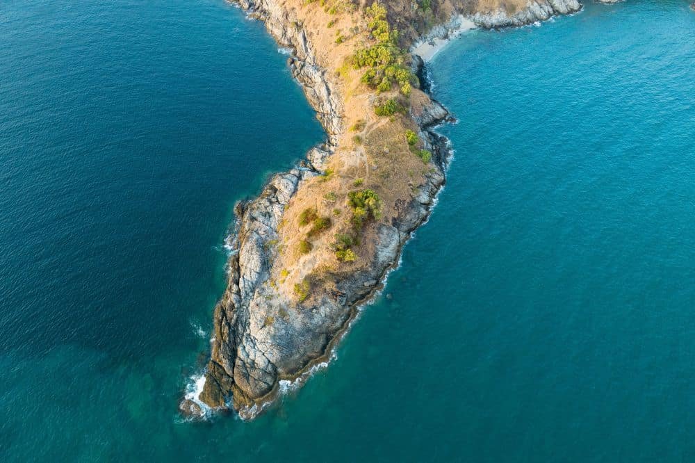 Aerial view of Laem Phromthep, a rocky, narrow peninsula jutting into blue-green ocean waters. The land is rugged with sparse vegetation on the top ridges, transitioning to rocky cliffs that lead down to the sea. The surrounding ocean is calm with gentle waves lapping against the shore.