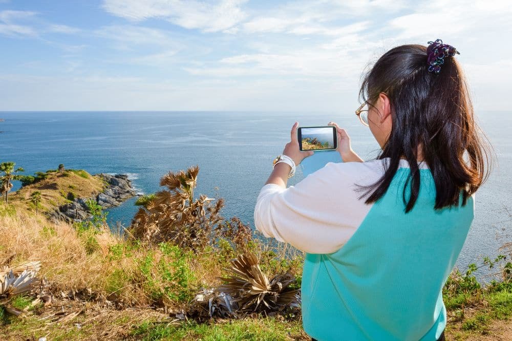 A person with long dark hair, wearing glasses and a turquoise and white top, is holding up a camera or phone to capture the breathtaking view from Laem Phromthep Sunset Viewpoint. The scene includes a vast blue ocean, a clear sky with wispy clouds, and cliffs adorned with greenery and dried plants.